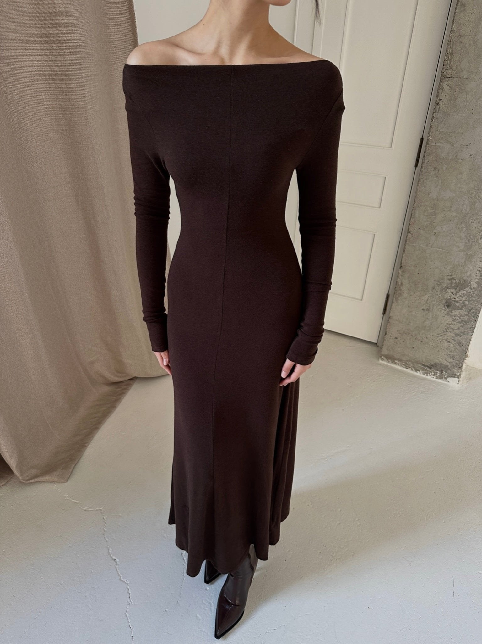 A person stands indoors wearing the Sienna Maxi Dress, a long, form-fitting black dress crafted from high-quality fabric with long sleeves, paired with black ankle boots. The backdrop comprises a beige curtain, a white door, and part of an exposed concrete wall, creating an overall minimalist setting.