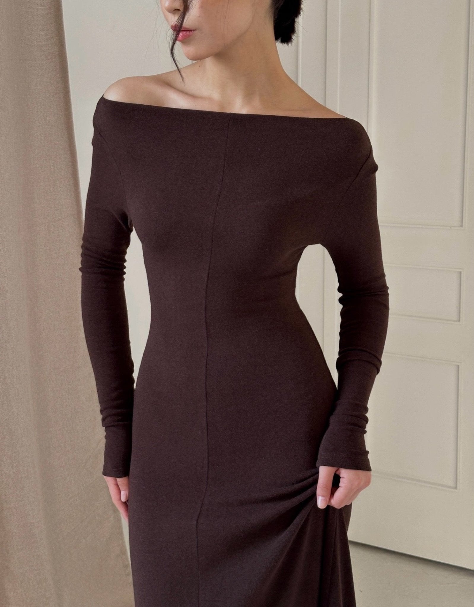 A person wearing a Sienna Maxi Dress stands indoors. The long, fitted brown dress has long sleeves and clings to their silhouette. Their hair is pulled back, and they look slightly downward while gently holding the dress at the side.