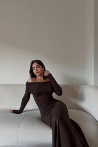 A woman with long black hair, wearing the Sienna Maxi Dress made from high-quality fabric, is sitting on a white sofa. She rests her left hand on the sofa while her right hand touches her hair, gazing slightly downward with a calm expression.