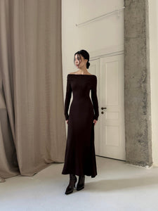 A woman stands in a minimalist room with white walls and a beige curtain, wearing a long Sienna Maxi Dress and black boots. Her hair is styled in an updo, and she looks to the side with a calm expression. A white door is slightly ajar behind her.