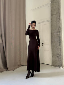 A person stands indoors wearing the Sienna maxi dress paired with brown lace-up boots. With one hand touching their head, they are positioned in front of a white door and a large beige curtain. The versatile dress, designed with high-quality fabric, complements the minimalist, modern aesthetic perfectly.