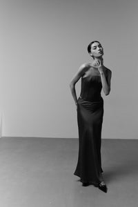 A black and white photograph captures a woman standing in a minimalist setting. She is wearing an elegant, long strapless dress that features the luxurious **Silky maxi skirt**. One hand delicately touches her neck while the other rests on her hip. Her hair is slicked back, and she gazes upwards.