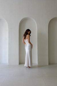 A woman with long brown hair stands with her back to the camera, showcasing the elegance of a Regina Corset Set with a maxi skirt. The fitted bodice and flowing skirt exude luxury, reminiscent of an opulent satin gown. She stands in front of an archway in a minimalist, light-colored room.