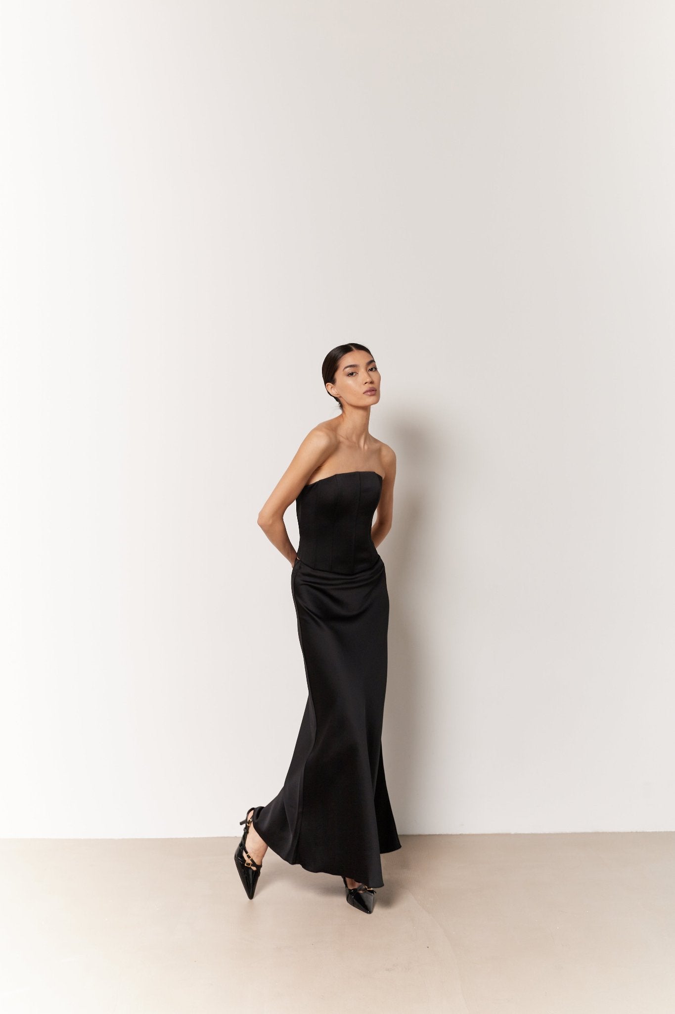 A woman dressed in a sleek, strapless gown featuring the Silky maxi skirt in black stands against a plain light-colored wall. She is wearing black pointed-toe shoes and has her hair pulled back neatly. Her arms are positioned back, and she is looking slightly up and to the side.