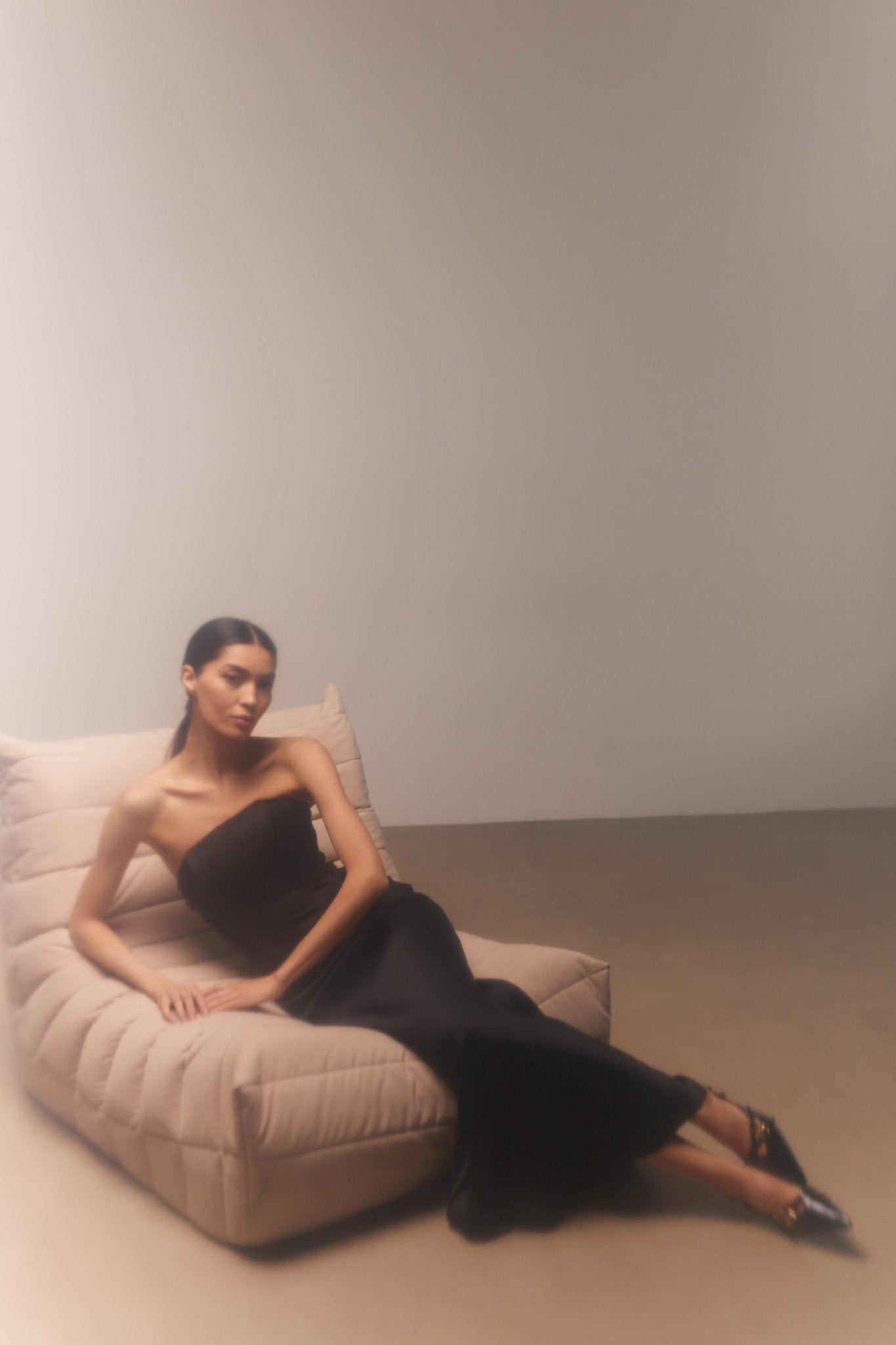 A person with long, dark hair is reclining on a beige, modern, cushioned chaise lounge. They are wearing a strapless black gown paired with a luxurious Silky maxi skirt and black pointed-toe high heels. The minimalistic background in neutral tones accentuates the elegance of their outfit.