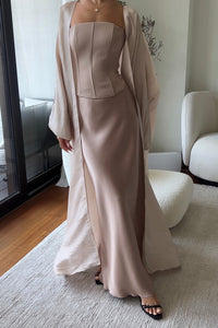 A person is standing indoors wearing a tan strapless dress with a fitted bodice and a flowing open-front overcoat. They have on black strappy heels. The room, adorned with wooden floors, a white cushioned chair, and a light-colored rug, perfectly complements their Silky maxi skirt.