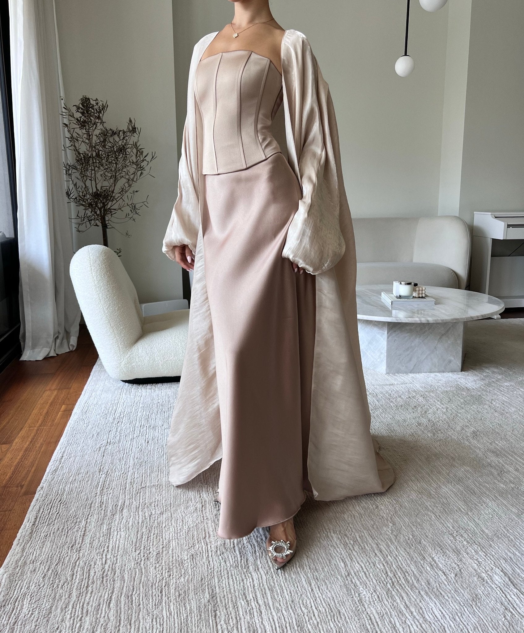 A person stands in a minimalist living room wearing an elegant blush pink strapless gown with a luxury Silky Maxi Skirt and a draped cardigan. The outfit is complemented by embellished heels. The room features modern furniture, including a white armchair, a light rug, and a round coffee table.