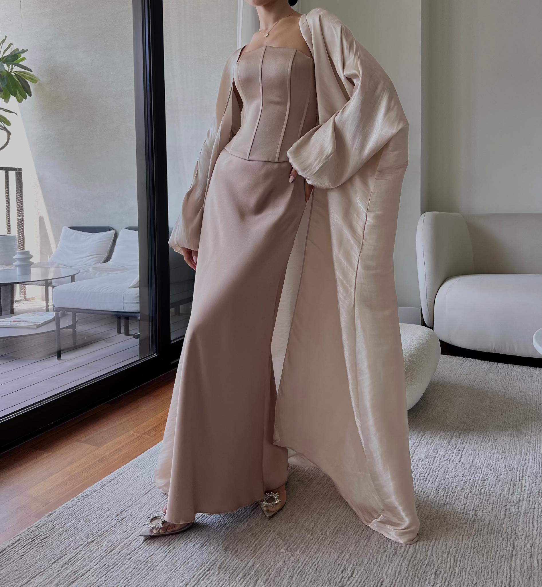 A person is standing indoors wearing a sophisticated light pink floor-length gown with a corset bodice. Complemented by a matching, draped overcoat, they pose with one hand on their hip. The room features modern, minimalist furniture and large windows. Consider pairing it with our Silky maxi skirt for added elegance.