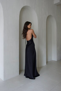 A woman wearing a sleek, backless Solange dress stands in an arched alcove. She is facing sideways, revealing her bare back with her hair cascading down. The background features smooth, white walls with arched cutouts, creating a serene, minimalist effect.