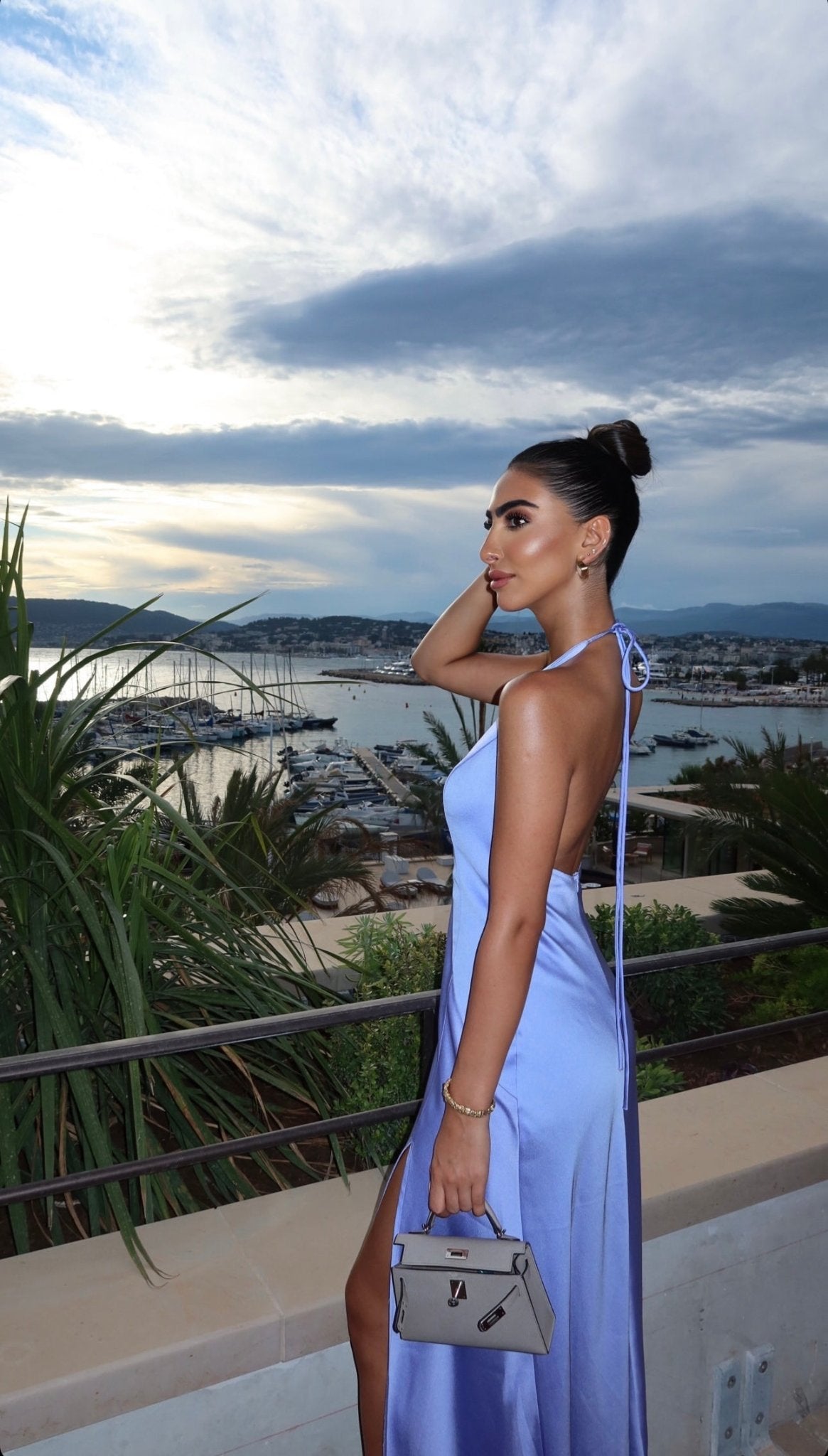 A woman in a light blue Solange Dress stands on a terrace overlooking a marina filled with boats. Holding a small handbag, she poses gracefully with one hand touching her hair, the silky fabric catching the light beautifully. The sky is partly cloudy, and it appears to be either sunset or sunrise.