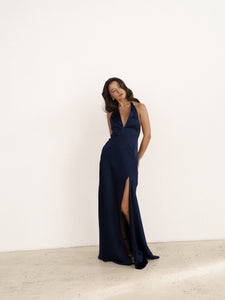 Against a plain wall, a woman confidently poses in a navy blue, sleeveless Solange Dress. The deep V-neck and high slit of the dress reveal one leg, while its silky fabric accentuates her elegant look. The backless design adds an extra touch of allure. With her hair styled in loose waves and one hand on her hip, she exudes sophistication.