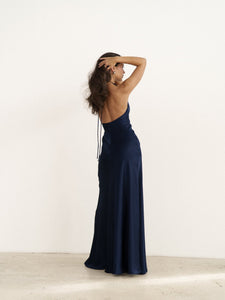 A woman stands against a plain white background, wearing a navy blue Solange Dress featuring a halter neck and long, silky fabric that drapes elegantly. Her long, wavy hair cascades down as she holds one hand to her head, looking away from the camera.