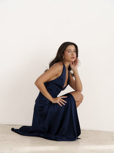 A woman with long, dark hair sits pensively on the ground in a silky, dark blue Solange dress. She rests her chin on her hand and gazes slightly off to the side against a plain white background.