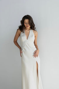 A woman stands against a white background, gently touching her hip and looking downward. Her long, wavy hair cascades over her shoulders. She is wearing the Solange dress, a sleeveless white satin garment featuring a deep V-neckline, a thigh-high slit on the left side, and silky fabric that glimmers softly in the light.