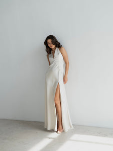 A woman models a long, flowing Solange Dress featuring a deep V-neckline and high slit as she stands against a plain white wall. The backless dress, made of silky fabric, cascades elegantly. With long, wavy brown hair and standing barefoot on a sunlit, light-colored floor, she gazes down with a gentle expression.