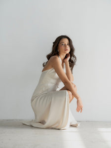 A woman with long, wavy hair is wearing the elegant Solange dress and sitting on her heels against a plain white background. She appears contemplative, with her head slightly tilted upward and her hands resting on her knees.