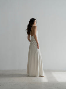 A woman with long, dark hair stands in a minimalist room with white walls and a light floor. She is wearing a sleeveless, white Solange dress made of silky fabric that glimmers in the sunlight streaming through an unseen window. Shown in side profile, she looks slightly to her left.