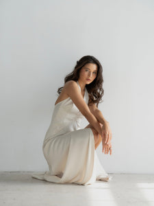 A woman with long, wavy hair wearing a Solange dress is sitting on the floor against a plain white background. She is looking straight ahead with a neutral expression, one knee bent and the other leg extended slightly to the side.