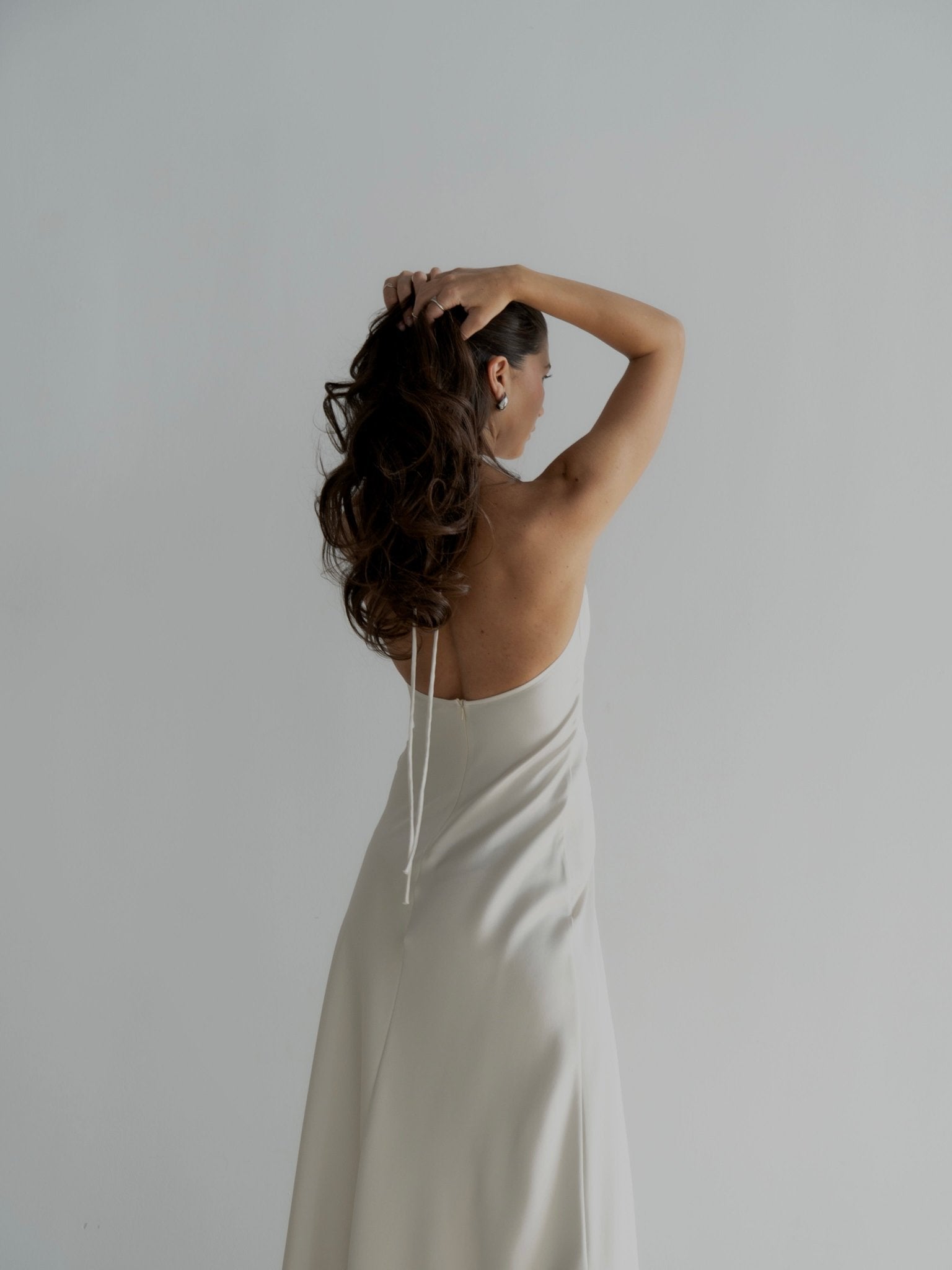 A woman with long, wavy hair stands with her back to the camera, showcasing a backless Solange Dress. The dress is crafted from silky fabric and drapes elegantly. She raises one arm to rest her hand on her head, while the other arm hangs casually by her side against a plain white background.