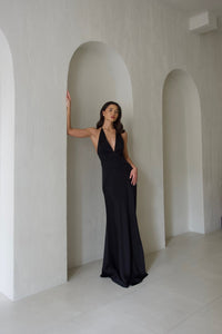 A woman with long dark hair stands elegantly in a minimalist, arched hallway. She is wearing a sophisticated black Solange dress that features a silky fabric, deep V-neck, and backless design. Resting one arm against the arch and looking confidently forward, she exudes refinement against the light-colored walls and floor tiles.