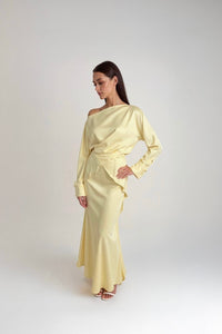 A woman standing against a white background wearing a light yellow outfit with balloon sleeves and a flowing skirt.