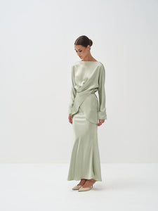A woman standing against a white background wearing a light green outfit with balloon sleeves and a flowing skirt.
