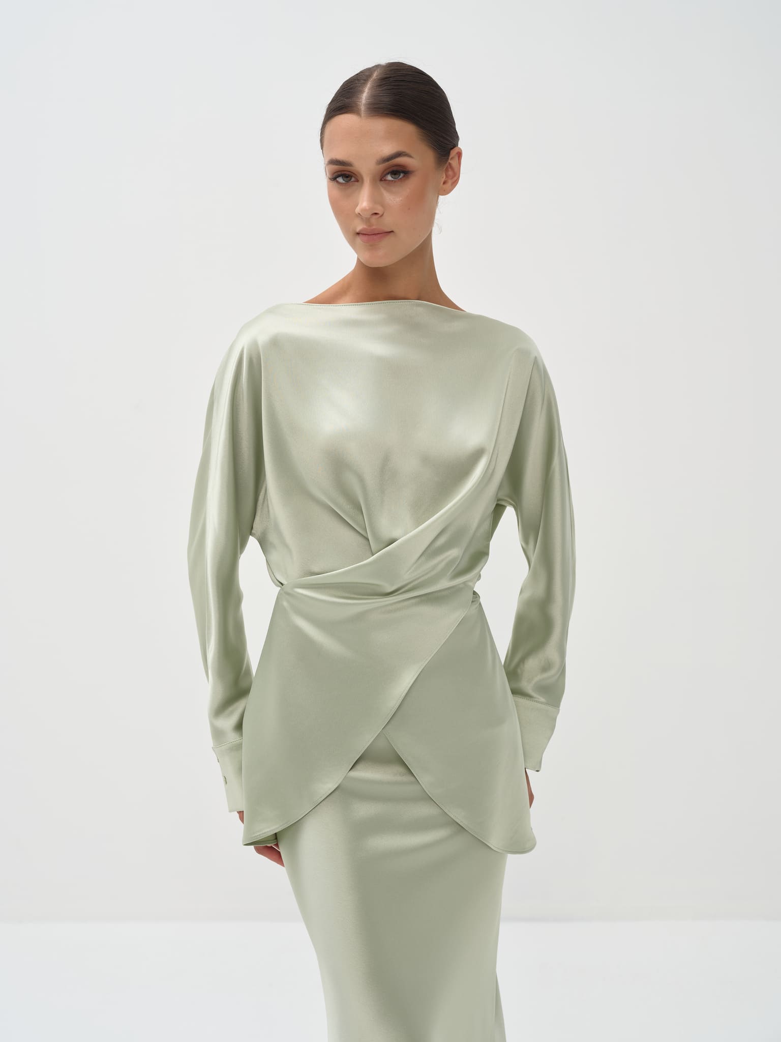 A woman standing against a white background wearing a light green outfit with balloon sleeves and a flowing skirt.