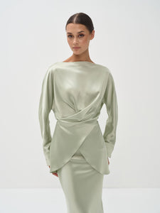 A woman standing against a white background wearing a light green outfit with balloon sleeves and a flowing skirt.