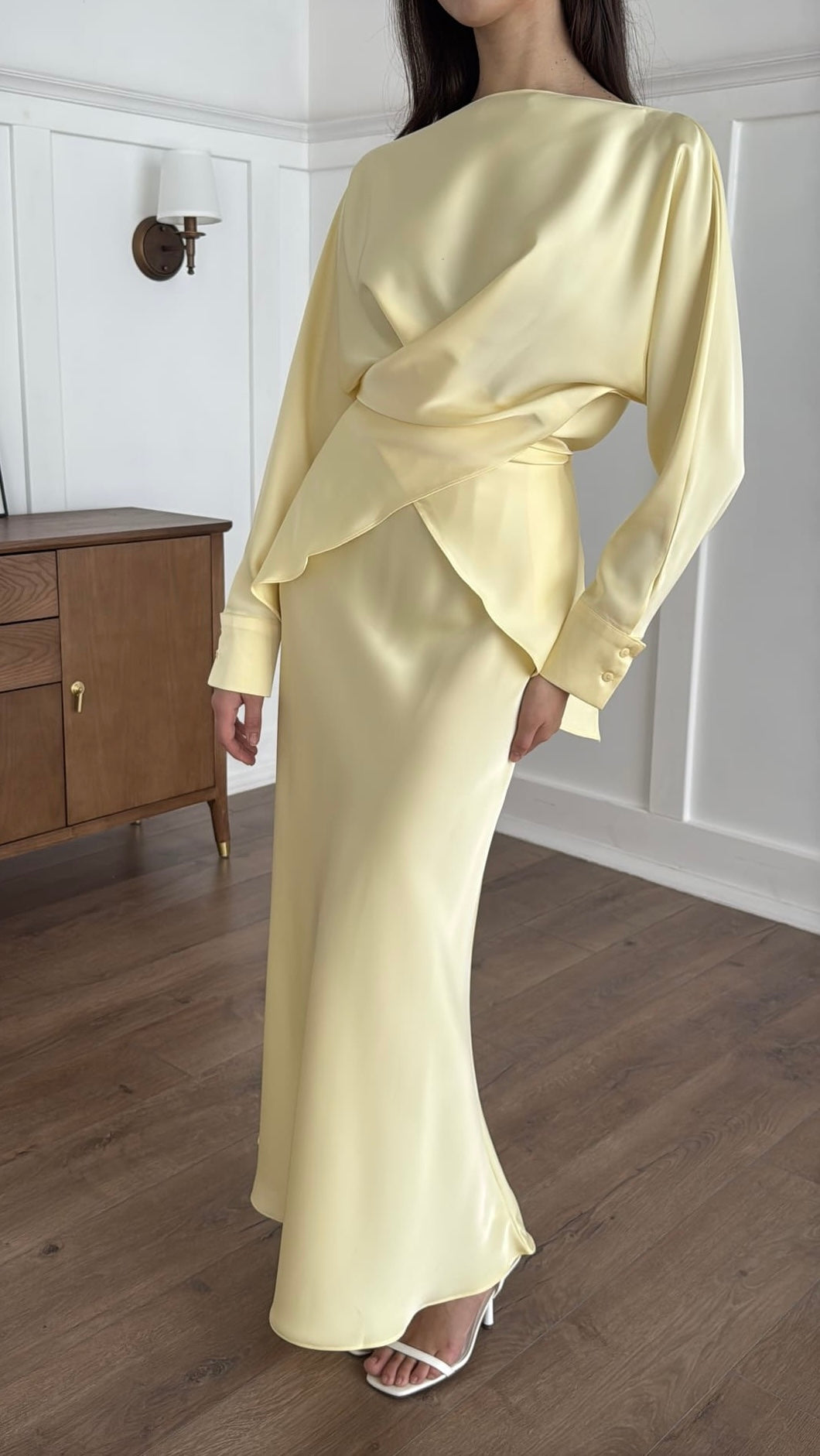 Person wearing a light yellow outfit standing in a room with wooden flooring and a white wall.