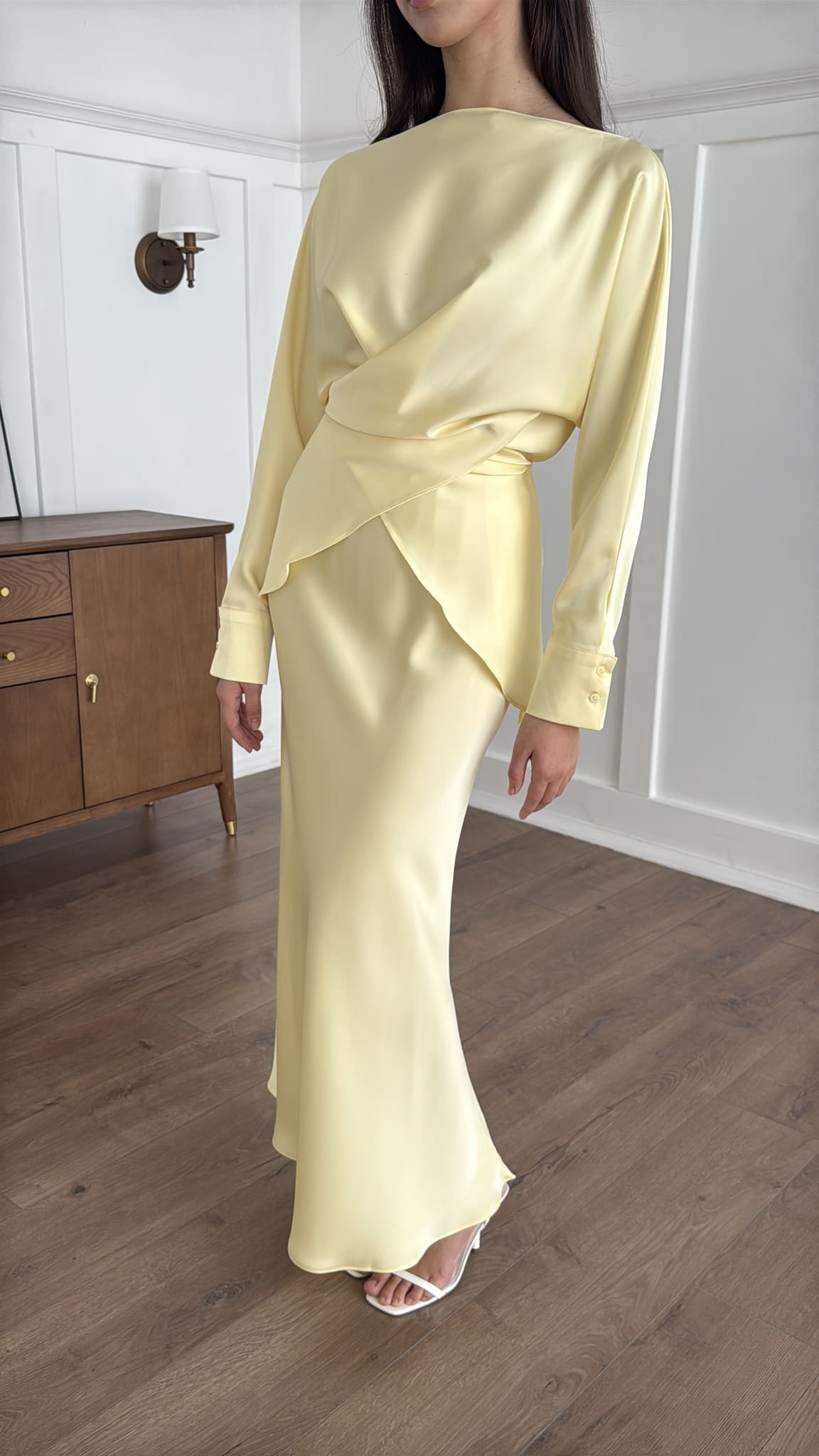 Woman wearing a light yellow outfit standing in a room with wooden flooring and white walls.
