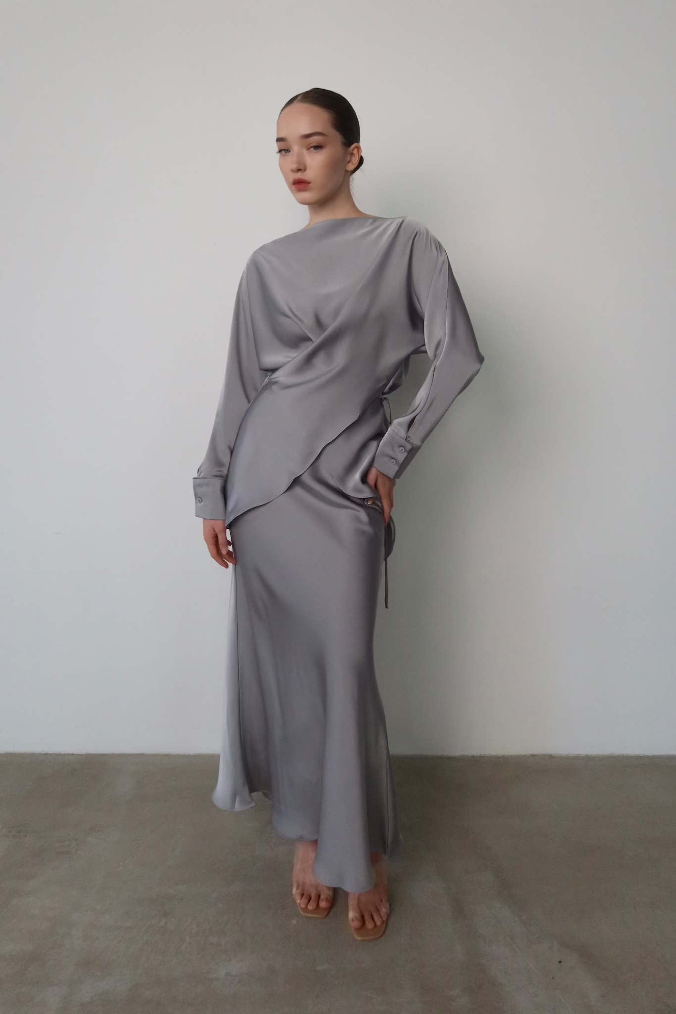 A person stands against a plain white wall, dressed in the sophisticated Tessa set—a sleek, long-sleeve light gray satin dress that drapes elegantly. They exude modern sophistication while posing with one hand on their hip and their other arm relaxed by their side. Their hair is pulled back, and they wear neutral makeup.