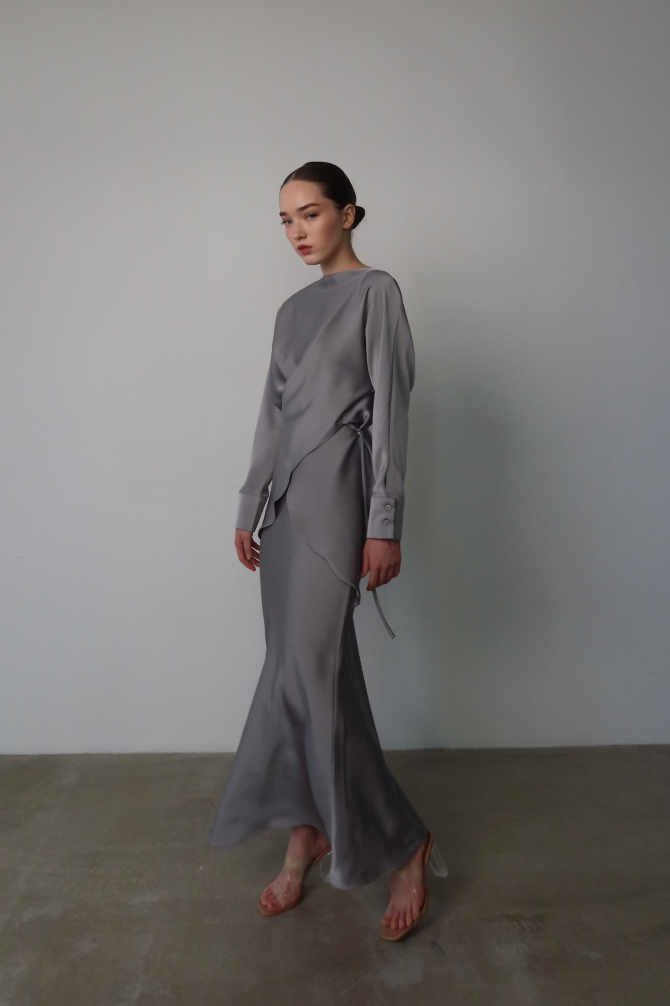 A person stands against a plain background wearing the Tessa set: a long, grey, flowy dress. Their hair is pulled back into a sleek bun, and they are wearing high-heeled clear sandals. The dress features long sleeves and an asymmetrical design, adding modern sophistication to the overall look.