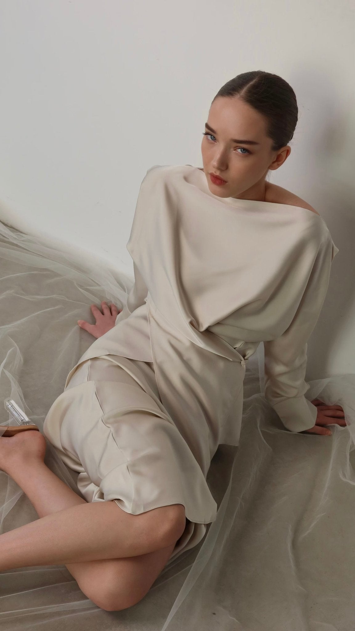 A woman with slicked-back hair is sitting on the floor against a white wall. She is wearing the Tessa set, an elegant, light-colored ensemble featuring long sleeves and draped fabric, exuding modern sophistication. Her legs are bent, one tucked under the other. She looks directly at the camera with a serious expression.