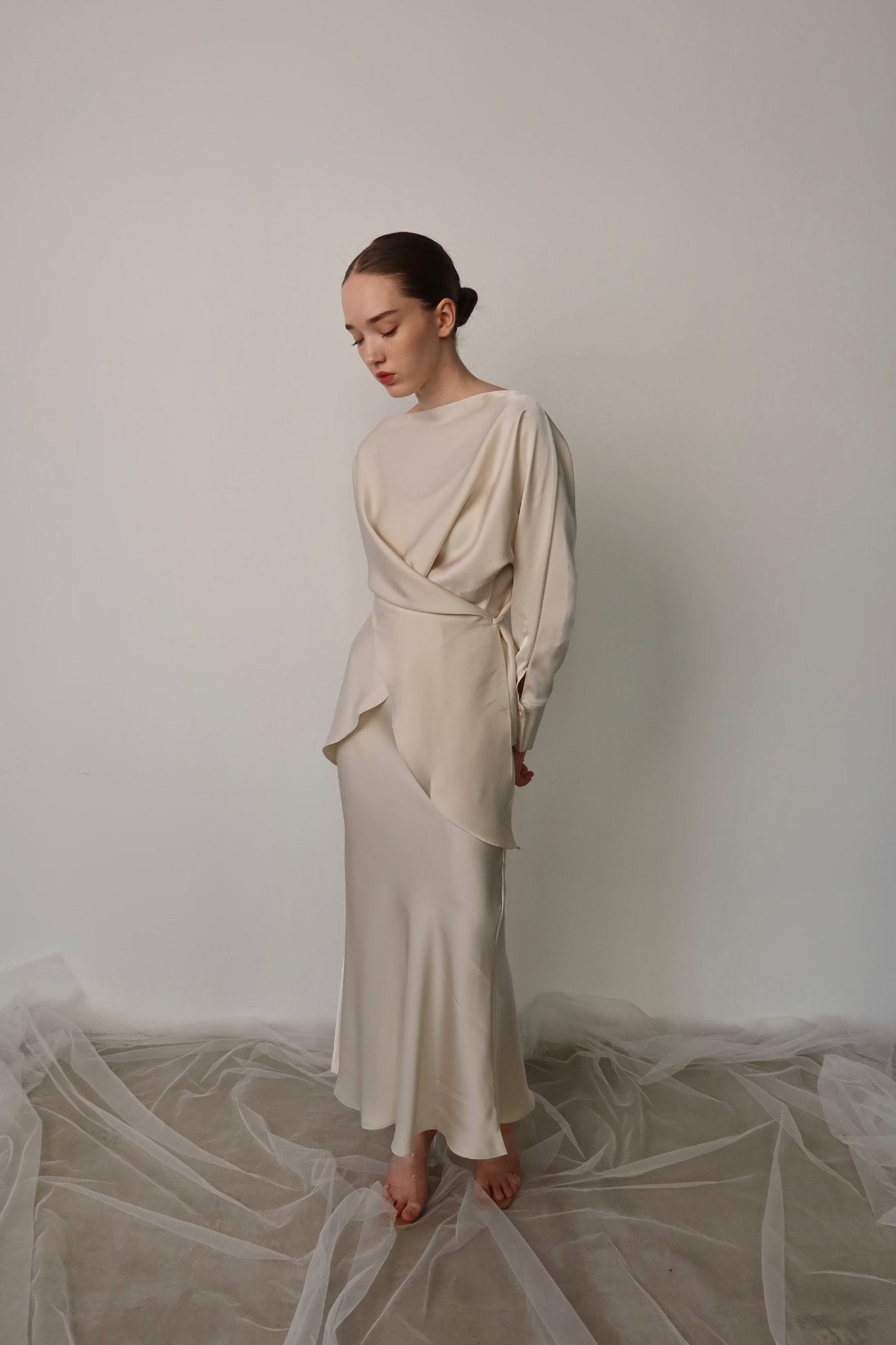 A woman stands barefoot on a taupe floor draped with sheer fabric, against a plain light gray wall. She is wearing the Tessa set, a long, elegant cream-colored dress with asymmetric draping that exudes timeless style. Her hair is pulled back into a sleek bun as she gazes downward with a calm expression.