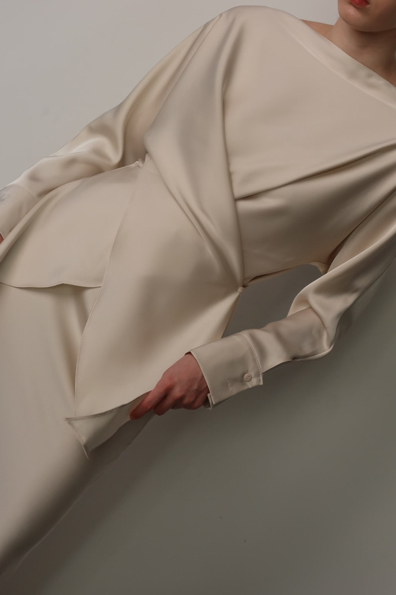 Close-up of a person wearing the Tessa set, a cream-colored satin dress with long sleeves and an asymmetrical, layered design. This luxurious ensemble features a loose-fit top and a fitted skirt, with the person’s right hand lightly holding a layer of the fabric near the hip.