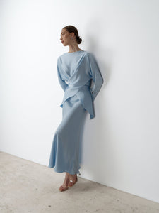 A woman wearing the Tessa set, a light blue, long-sleeved, draped dress, is leaning against a white wall. Exuding modern sophistication, her hair is styled in a sleek bun as she gazes off to the side. She completes her luxurious ensemble with clear, open-toe heels and stands on a light-colored stone floor.