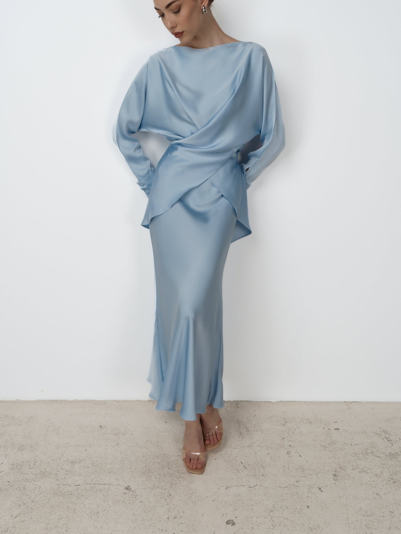 A person wearing the Tessa set, featuring a light blue, long-sleeved asymmetrical dress, stands against a plain white background. The luxurious ensemble drapes elegantly with a flowing design and is complemented by clear high-heeled sandals. Exuding modern sophistication, the person gazes downwards.