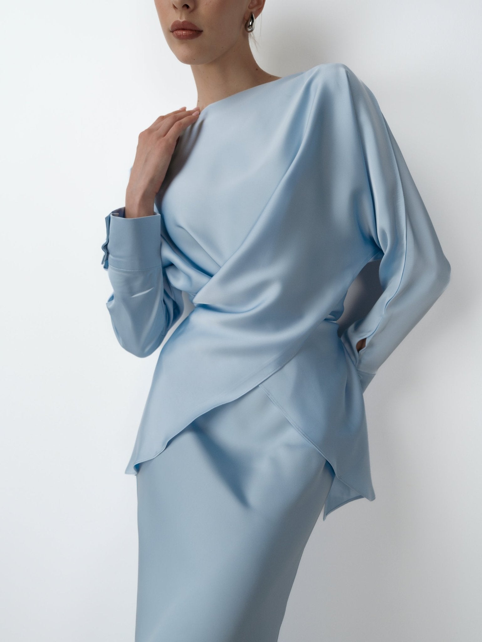 A person wearing the elegant Tessa set, a light blue satin-like ensemble, poses against a plain white background. The outfit features long sleeves and an asymmetrical design with draped fabric, creating a timeless style. The person's hand rests gently on their shoulder, exuding modern sophistication.