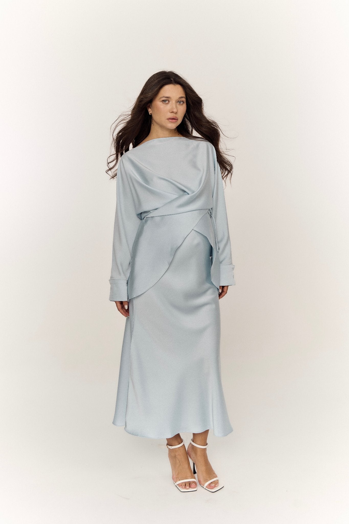 A woman with long, dark hair stands against a plain background, exuding modern sophistication in the light blue Tessa set. This ensemble features a long-sleeved dress with a layered design and a mid-calf length skirt, perfectly completed by her white heeled sandals.