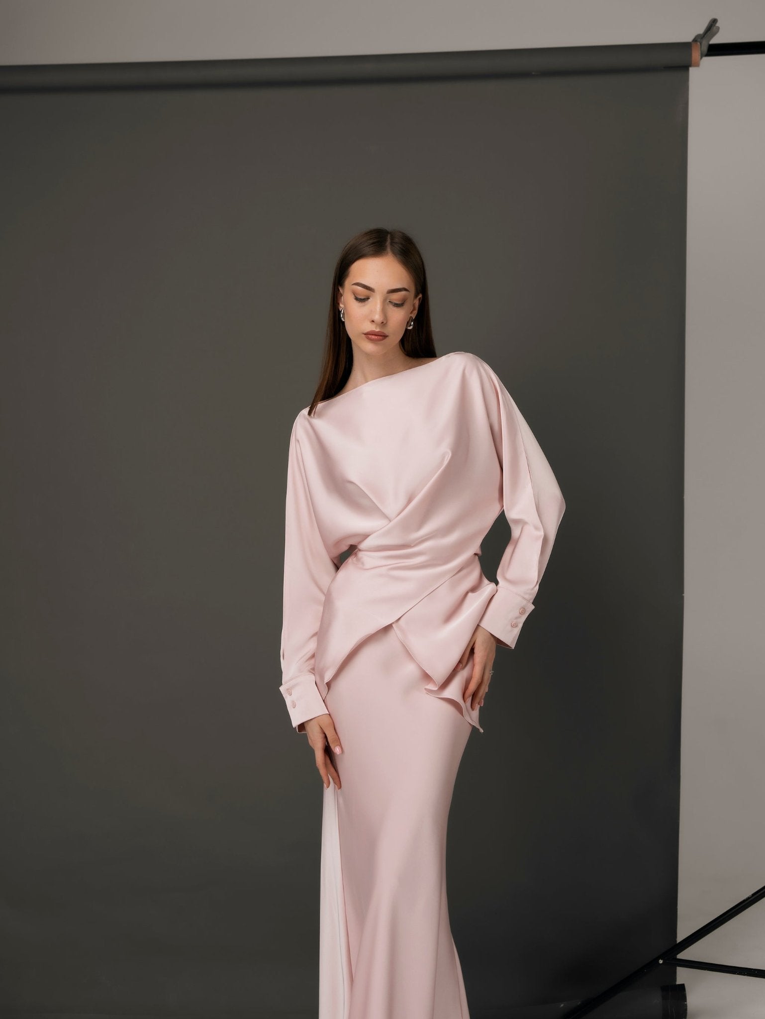 A woman with long, straight hair poses gracefully in the Tessa set, a soft pink, elegant dress with long sleeves and a wrap-around design. She stands against a plain dark backdrop, exuding timeless style and confidence through her luxurious ensemble.