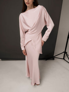 A person with long hair is wearing the elegant Tessa set in light pink, showcasing a draped design and posing against a neutral backdrop. This luxurious ensemble features long sleeves and a floor-length skirt. The person poses with one hand on their hip.