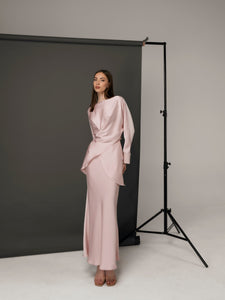 A woman stands in front of a dark gray backdrop, wearing the Tessa set—a silky, light pink top paired with a matching long skirt. She exudes modern sophistication with her straight, long brown hair and confident pose, one hand on her hip. Studio lighting and a backdrop stand are visible.