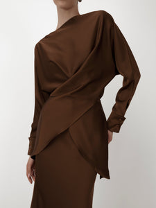 Person wearing the Tessa Set, showcasing a luxurious silky satin brown ensemble that includes a long-sleeve wrap blouse with elegant balloon sleeves and an asymmetrical design, paired perfectly with a matching brown skirt. The plain white background highlights the outfit's rich texture and drape.