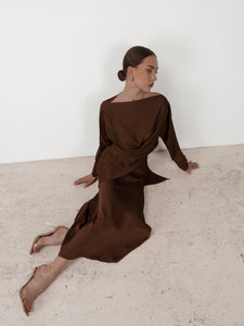 A woman wearing the Tessa set, a sleek, chocolate-brown gown made from luxurious silky satin and featuring elegant balloon sleeves, sits on the ground against a white wall. Her hair is styled in a neat bun, and she is wearing clear, open-toe heels. In a relaxed pose, one of her hands rests on the floor while the other is placed on her leg.