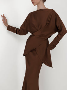 A person in the fashionable Tessa set, featuring a luxurious silky satin brown blouse with an asymmetric, draped design and elegant balloon sleeves. The back view reveals the intricate wrapping detail as the wearer’s left arm is raised, gently touching a wall against a plain white background.