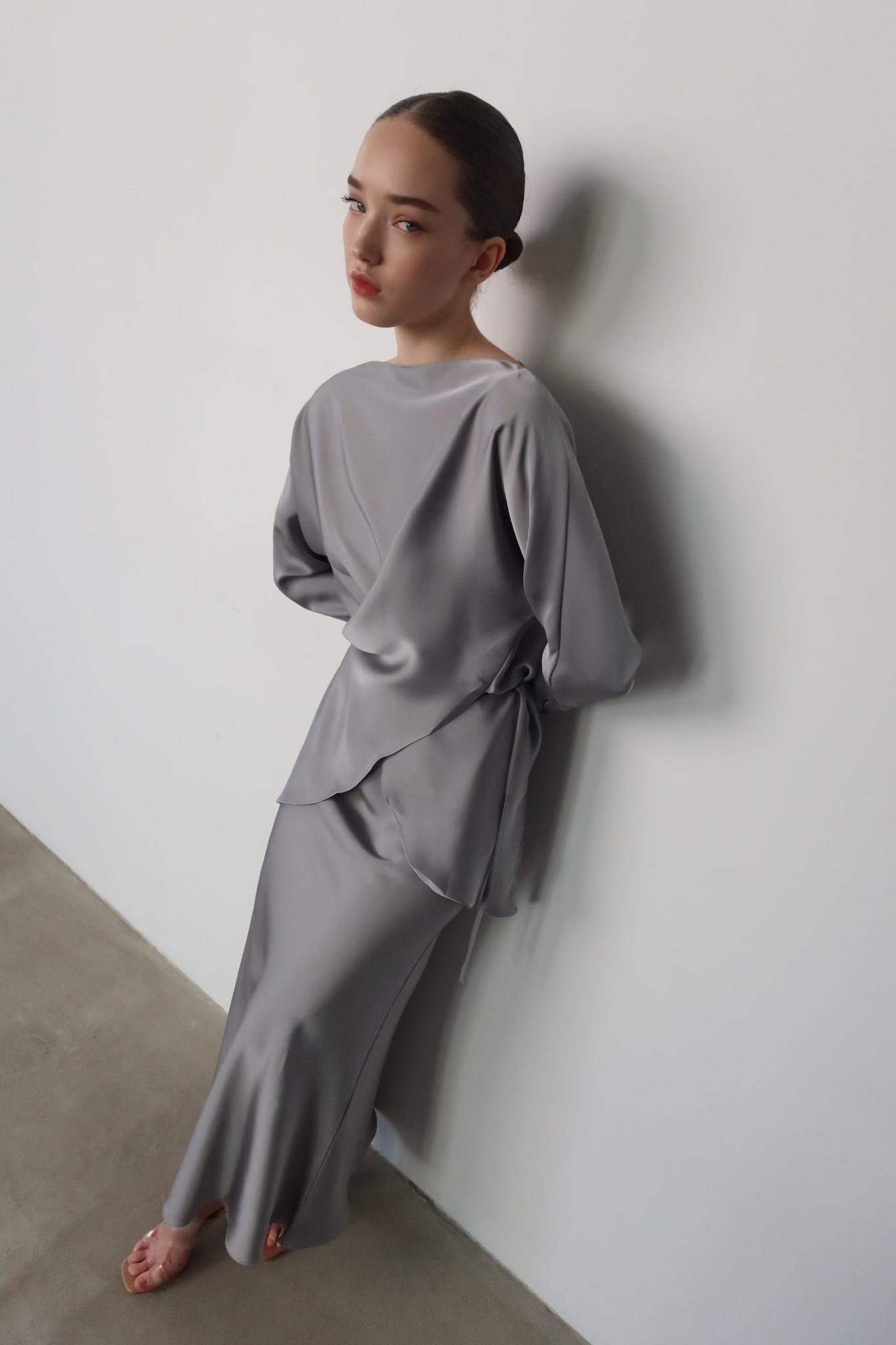 A woman with a neutral expression stands against a minimalist white wall. She is dressed in the Tessa set, featuring a flowing, light gray blouse with elegant balloon sleeves that are tied at the waist, paired with a matching long skirt. Her hair is neatly tied back, she is barefoot, and the setting appears to be indoors with natural lighting.