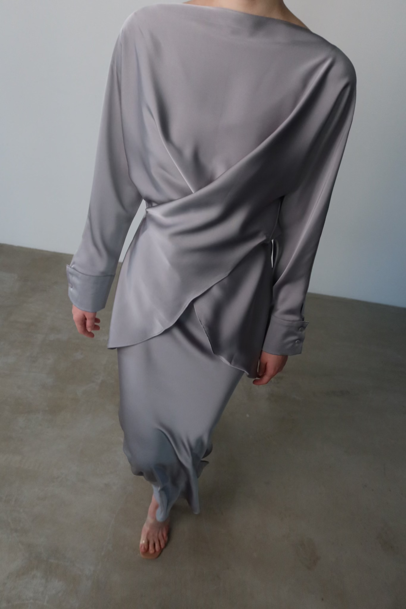 A person wearing the luxurious Tessa Set in a silky satin, silver-gray finish with elegant balloon sleeves and asymmetrical details, is walking barefoot on a smooth concrete floor. Captured from behind, the image beautifully showcases the flowing fabric and exquisite design of the dress.