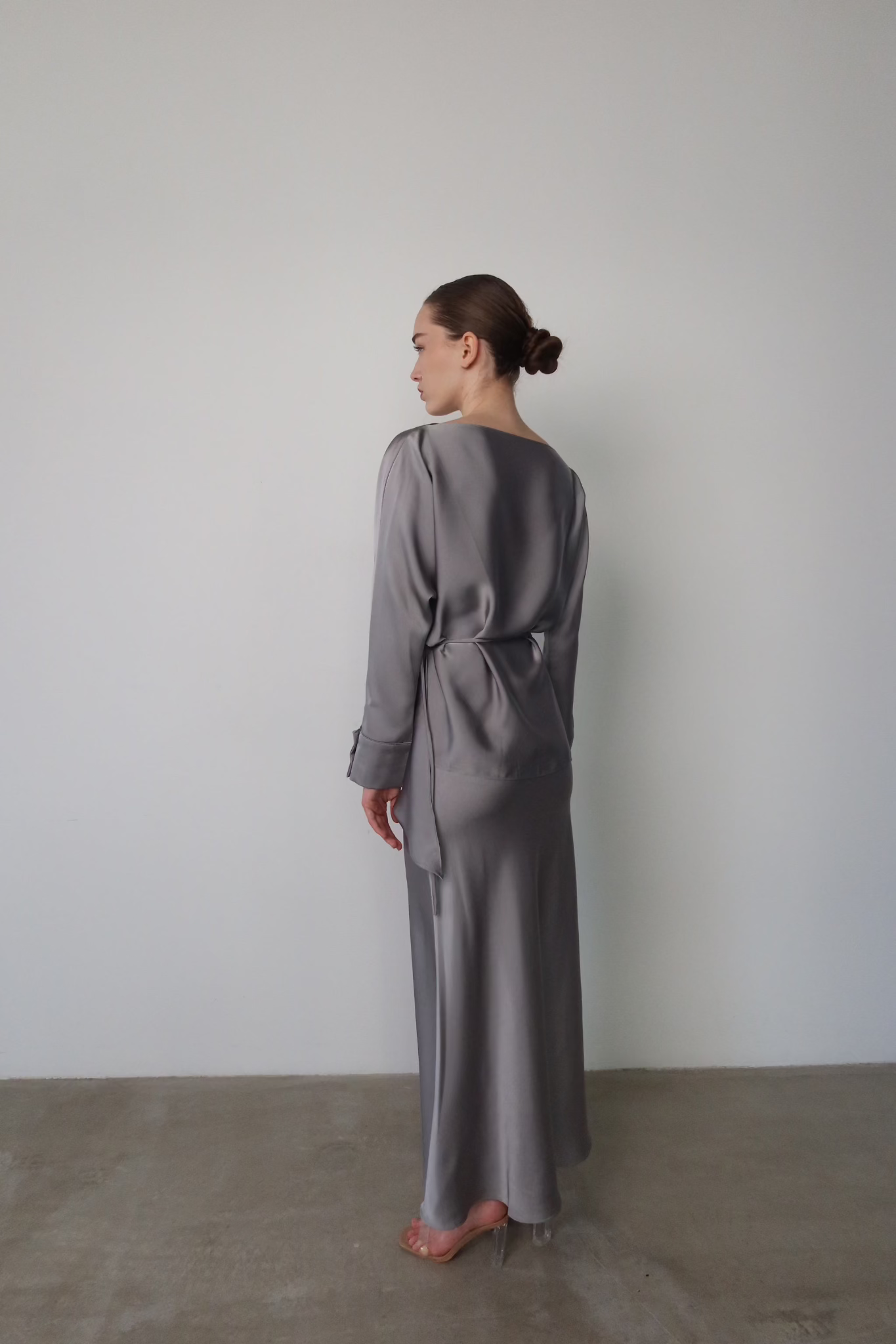 A model in profile view is showcasing the Tessa Set, an exquisite long dress crafted from luxurious silky satin featuring elegant balloon sleeves. Her hair is styled neatly in a bun. The backdrop is minimalist with a light-colored wall and concrete floor, enhancing the elegant ambiance.