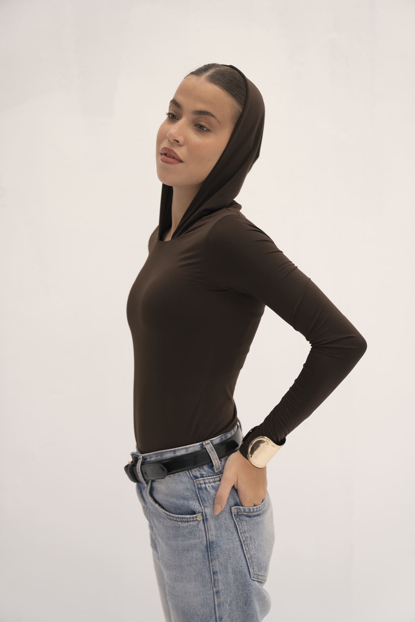 Wearing the Urban Drape Top, a woman in profile stands against a plain background. The sleek, long-sleeve, dark brown hooded top complements her light blue jeans and black belt. Her hair is pulled back as she rests her hand on her hip and gazes calmly ahead, perfectly capturing an urban drape aesthetic.