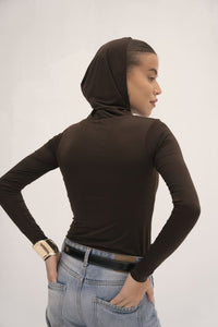 A woman facing away from the camera showcases versatile styling in an Urban Drape Top—black, long-sleeved with a hood—paired with jeans. Her hair is tied back, and she wears a gold bangle on her right wrist, making this contemporary piece stand out against the plain white backdrop.